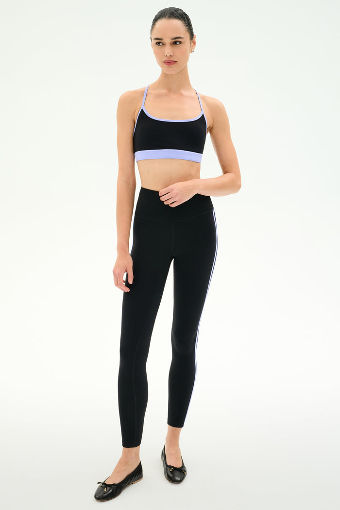 A woman stands against a white background wearing the SPLITS59 Emerson Airweight Bra in Black/Purple Haze, paired with high-waisted black nylon leggings featuring side stripes and black ballet flats.