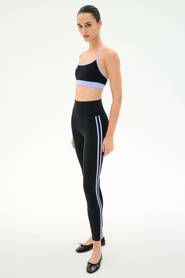 A woman stands on a white background wearing the SPLITS59 Emerson Airweight Bra in Black/Purple Haze, paired with high-waisted black leggings with light blue stripes and black ballet flats.