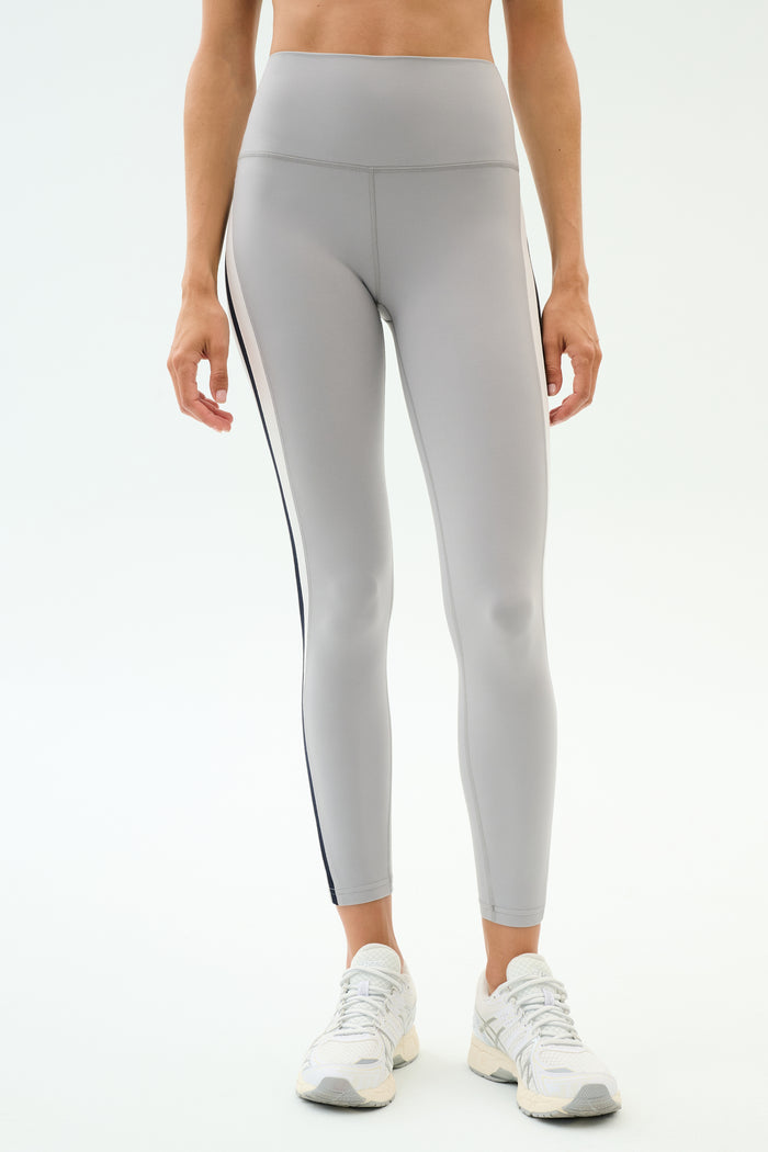 A person models SPLITS59 Emerson Airweight High Waist leggings in Dove Grey/Indigo—featuring a thin black side stripe and a sculpted fit—paired with white sneakers, perfect for hot yoga.