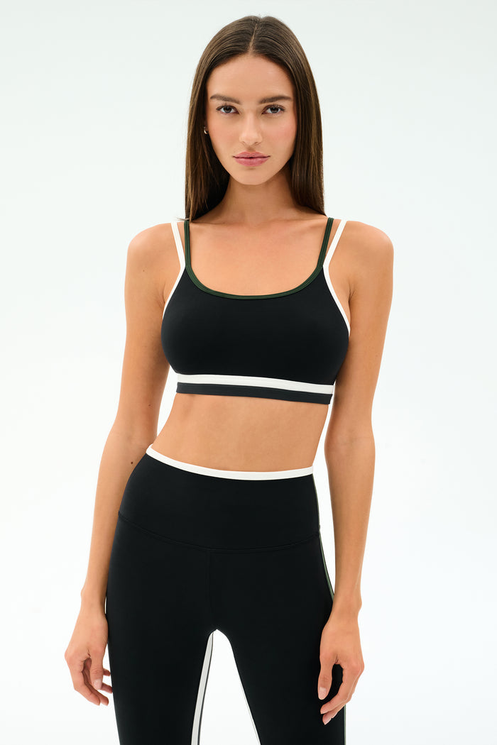 A woman stands against a plain background, wearing the SPLITS59 Eva Rigor Double Strap Bra in black with matching high-waisted leggings with white trim, looking directly at the camera.