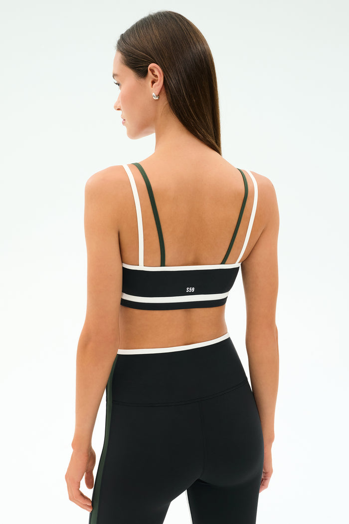 A woman with long brown hair faces away from the camera against a plain light background, wearing SPLITS59's Eva Rigor Double Strap Bra in Black/Military and black leggings.