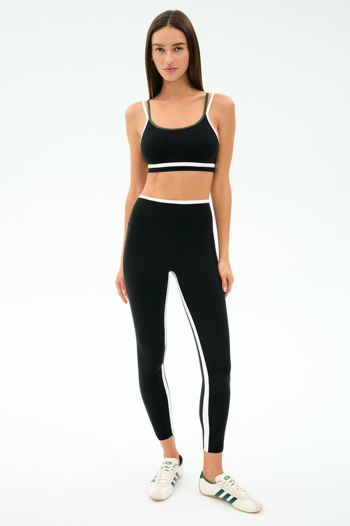 A woman faces forward wearing the SPLITS59 Eva Rigor Double Strap Bra in Black/Military, paired with matching black leggings featuring white stripes, and white sneakers with green accents.