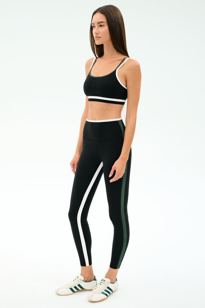 A woman stands in a studio wearing the SPLITS59 Eva Rigor Double Strap Bra in Black/Military, paired with matching black leggings featuring white and green stripes and white sneakers.