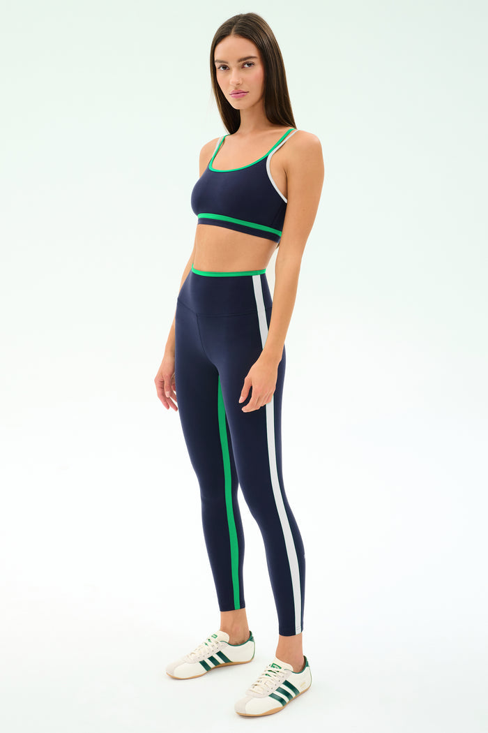A woman models the SPLITS59 Eva Rigor Double Strap Bra in Indigo/Spearmint, paired with leggings featuring green and white stripes and white sneakers, against a plain light background.