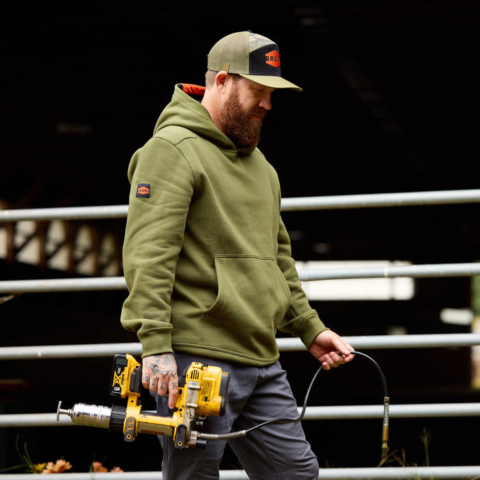 Man wearing flat brim work hat with built in pencil holder and BRUNT logo in army green camo carrying power tool * 0