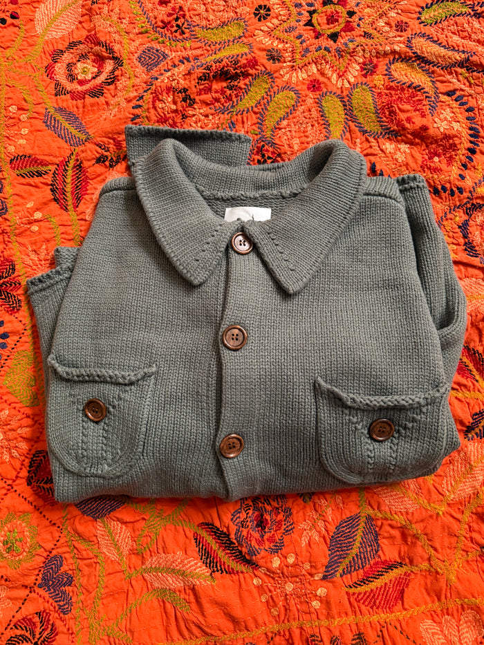 The Hunter Knit Collared Cardigan by FOUND, featuring wooden buttons and two front pockets, is laid on an orange floral-patterned fabric and offers a unisex relaxed fit for effortless style.