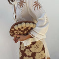 Jaden Beaded Clutch Bag with a brown and cream pattern and pom-pom trim, held by a model in a white embroidered shirt.