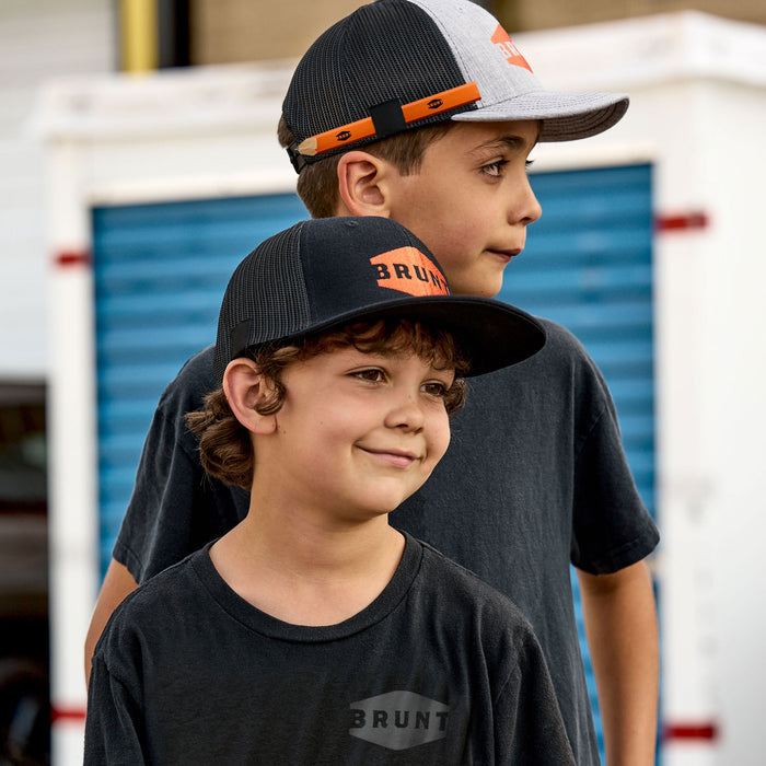 Two kids wearing kids curved brim work hat with built in pencil holder and BRUNT logo in grey and kids flat brim hat with built in pencil holder and BRUNT logo in black