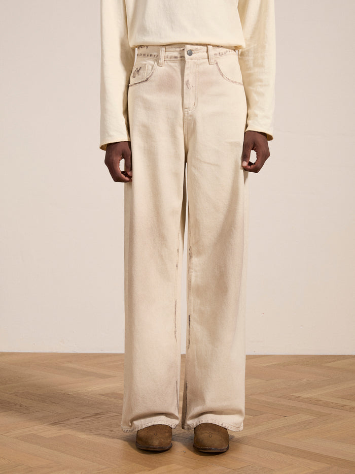 A person wearing a light beige long-sleeved shirt and the Lacy Baggy Jeans from FOUND in a dirty white wash, standing on a hardwood floor. Only the lower half of their body is visible.