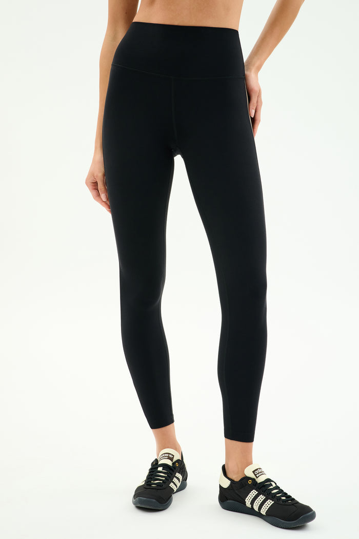 Wearing SPLITS59 Layla Airweight High Waist 7/8 in Black/Lentil and black sneakers with white stripes, standing against a light background—ideal for hot yoga or any workout.