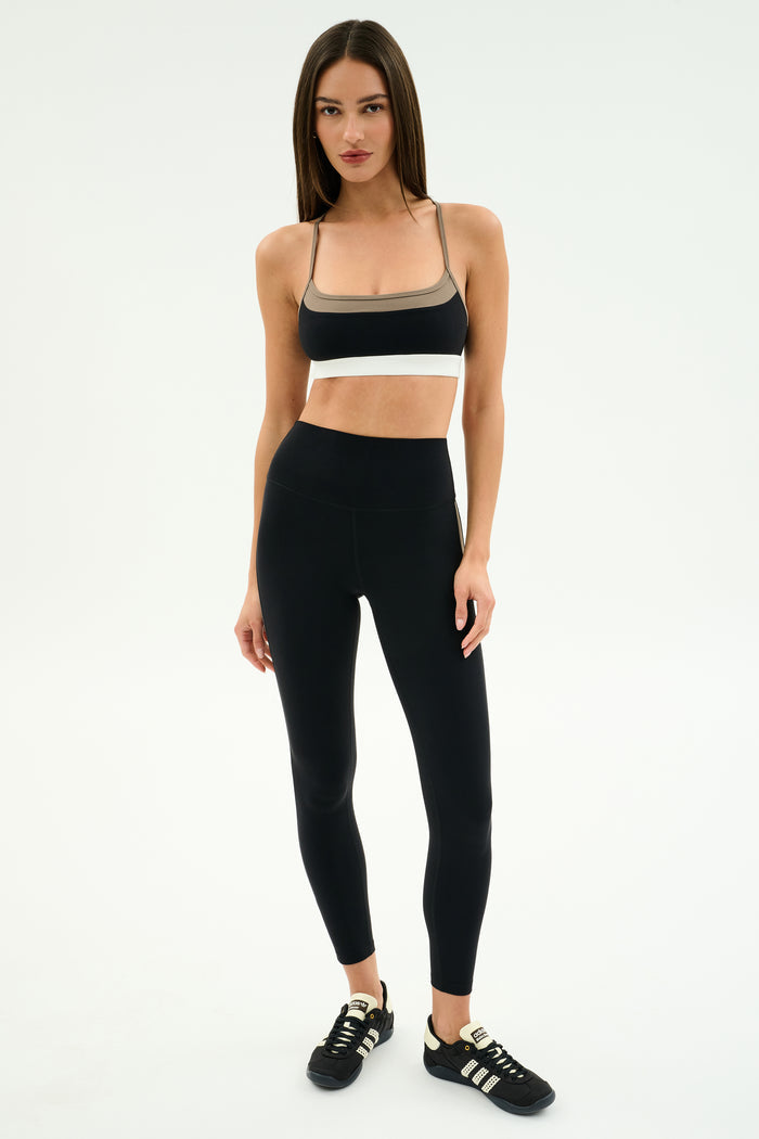 A woman stands against a plain background wearing SPLITS59 Layla Airweight High Waist 7/8 leggings in Black/Lentil with a black sports bra and black Adidas sneakers—ideal attire for hot yoga or gym sessions.