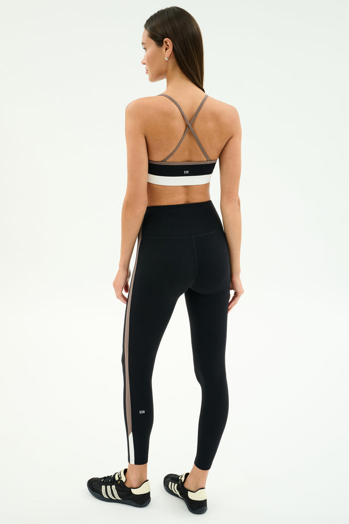 A woman stands facing away in SPLITS59's Layla Airweight High Waist 7/8 leggings in Black/Lentil, featuring side stripes and a cross-back sports bra—ideal for hot yoga—paired with black athletic shoes.