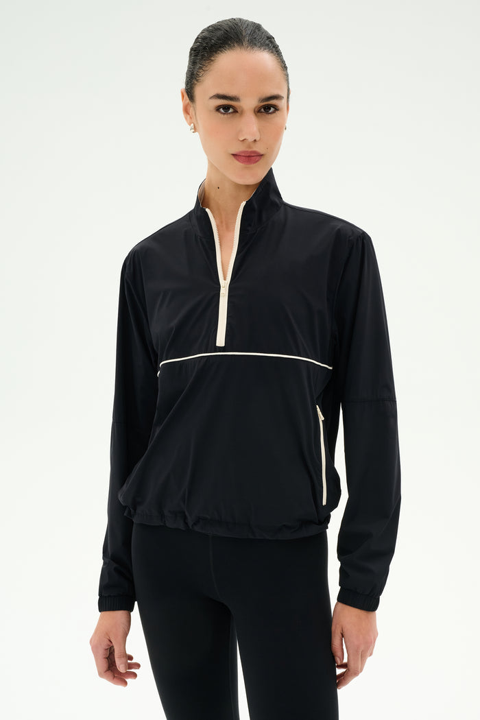 A woman wears the SPLITS59 Lyle Woven Nylon Pullover in Black/Creme with black spandex leggings, standing against a plain white background.