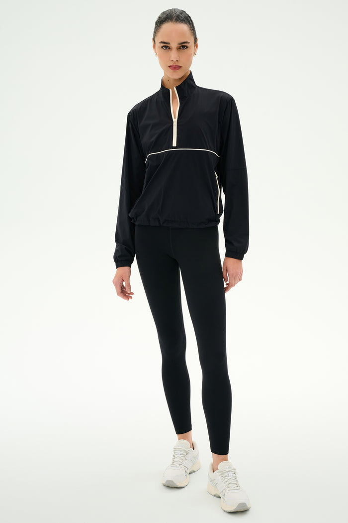 A person wearing the SPLITS59 Lyle Woven Nylon Pullover in Black/Creme, black spandex leggings, and white sneakers stands against a plain white background.