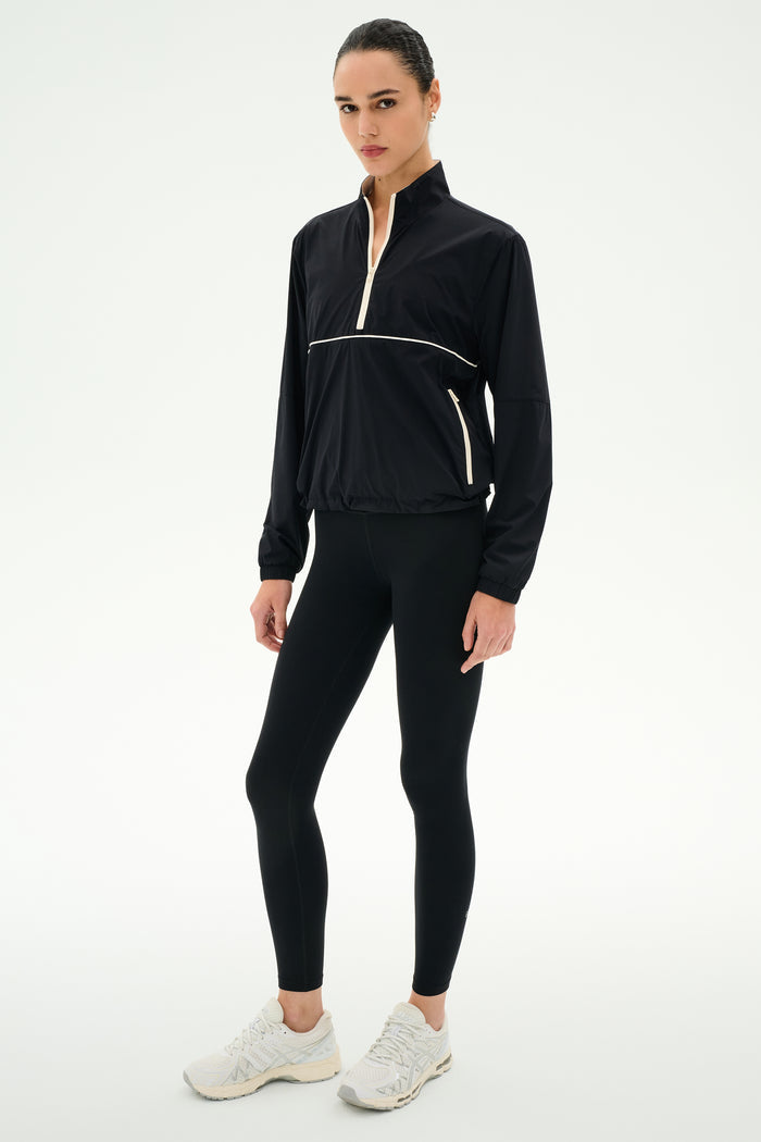 A woman wears the SPLITS59 Lyle Woven Nylon Pullover in Black/Creme, styled with spandex leggings and white sneakers, standing against a plain white background.