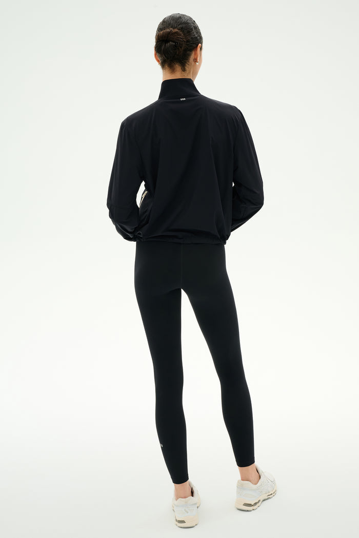 A person stands with their back to the camera, wearing the SPLITS59 Lyle Woven Nylon Pullover in Black/Creme, paired with black spandex leggings and white sneakers against a plain light background.