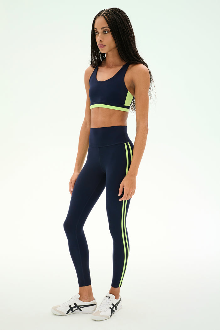 A woman is wearing the SPLITS59 Maci Airweight Bra in Indigo/Limeade, featuring neon yellow-green accents and crafted from a stretchy nylon spandex fabric, paired with white sneakers against a plain light background.