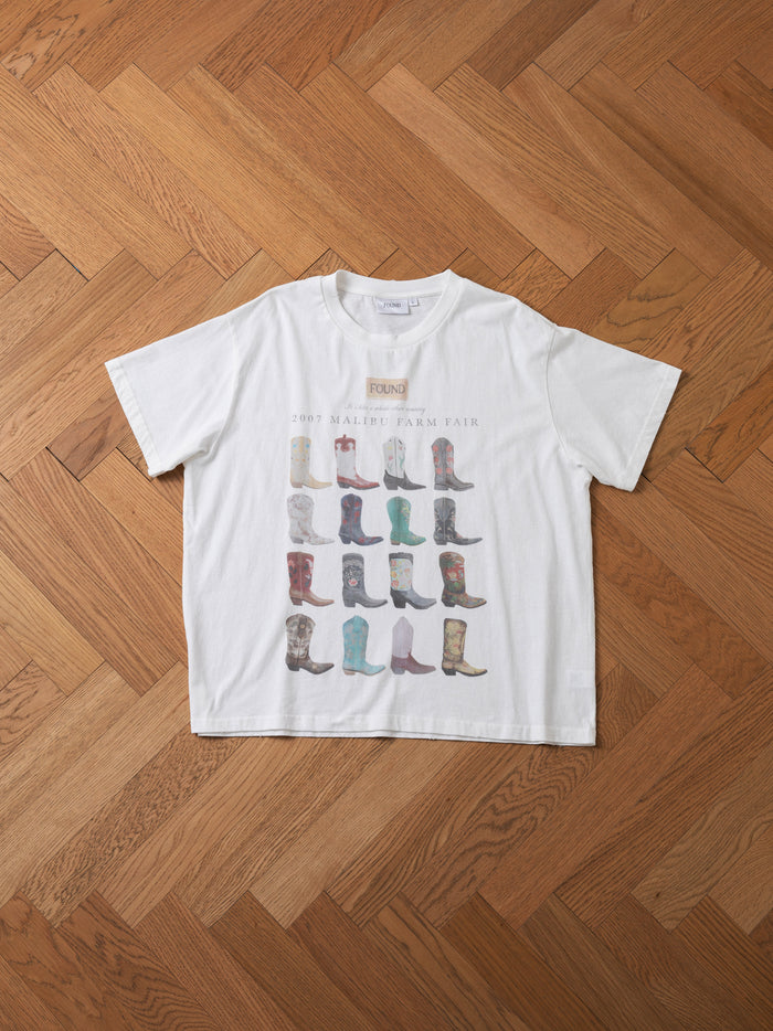 The FOUND Malibu Farm Fair Tee, made from 100% cotton, showcases the text "2007 Malibu Farm Fair" along with illustrations of various cowboy boots displayed on a wooden herringbone floor. This vintage farm fair t-shirt adds a timeless touch to your wardrobe.
