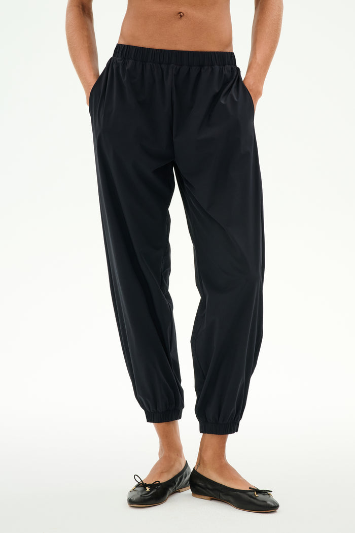 Model wears the SPLITS59 Megan Woven Nylon Jogger in black, featuring a cinched waistband and hands in pockets, paired with black ballet flats. Shown from chest down against a plain background.