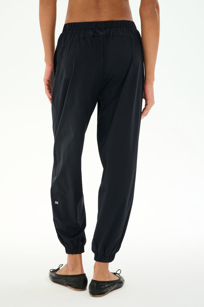 A person is seen from behind, standing against a white background, wearing SPLITS59 Megan Woven Nylon Jogger in black paired with black flat shoes.