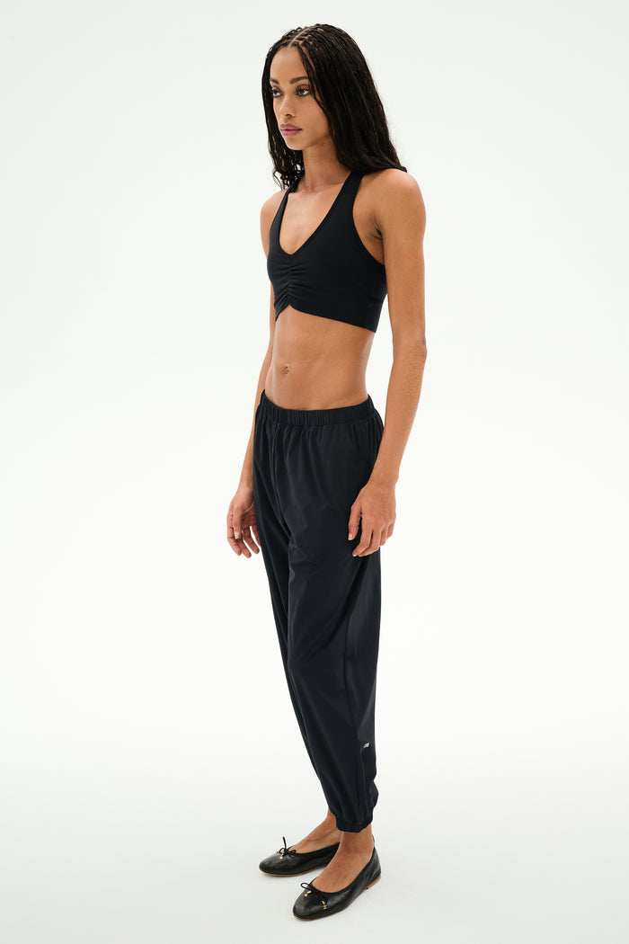 A person stands against a plain background wearing a black crop top, black SPLITS59 Megan Woven Nylon Jogger pants with a cinched waistband, and black ballet flats.