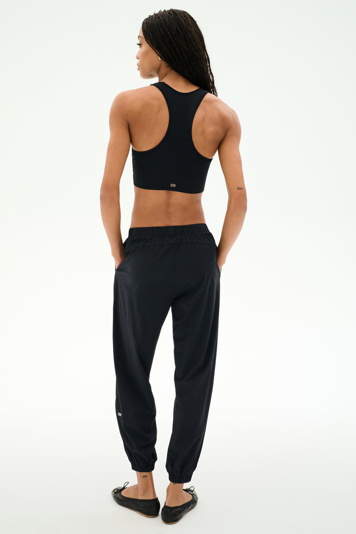 Person standing with their back to the camera, wearing SPLITS59’s Megan Woven Nylon Jogger - Black, a racerback sports top, and black shoes against a plain light background.