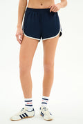 A person wears the SPLITS59 Megan Woven Nylon Run Short in Indigo/Creme, paired with white tube socks featuring red and blue stripes, and white sneakers with navy stripes, standing against a plain white background.