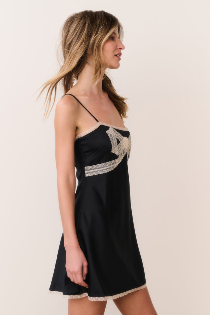Woman wearing a black satin dress with white lace trim and bow detail against a plain background