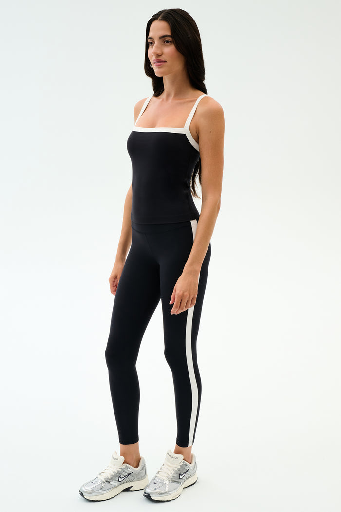 A woman wears the SPLITS59 Monah Rigor Tank in Black/White with matching black leggings featuring a white side stripe and silver athletic shoes—ideal gym attire—set against a plain white background.