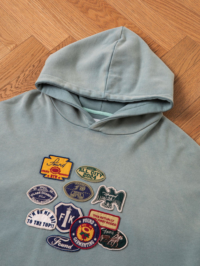 A Multi Patch Hoodie by FOUND, crafted from French Terry cotton, adorned with an array of colorful vintage-inspired embroidered patches on the front, is laid flat on a wooden herringbone parquet floor.