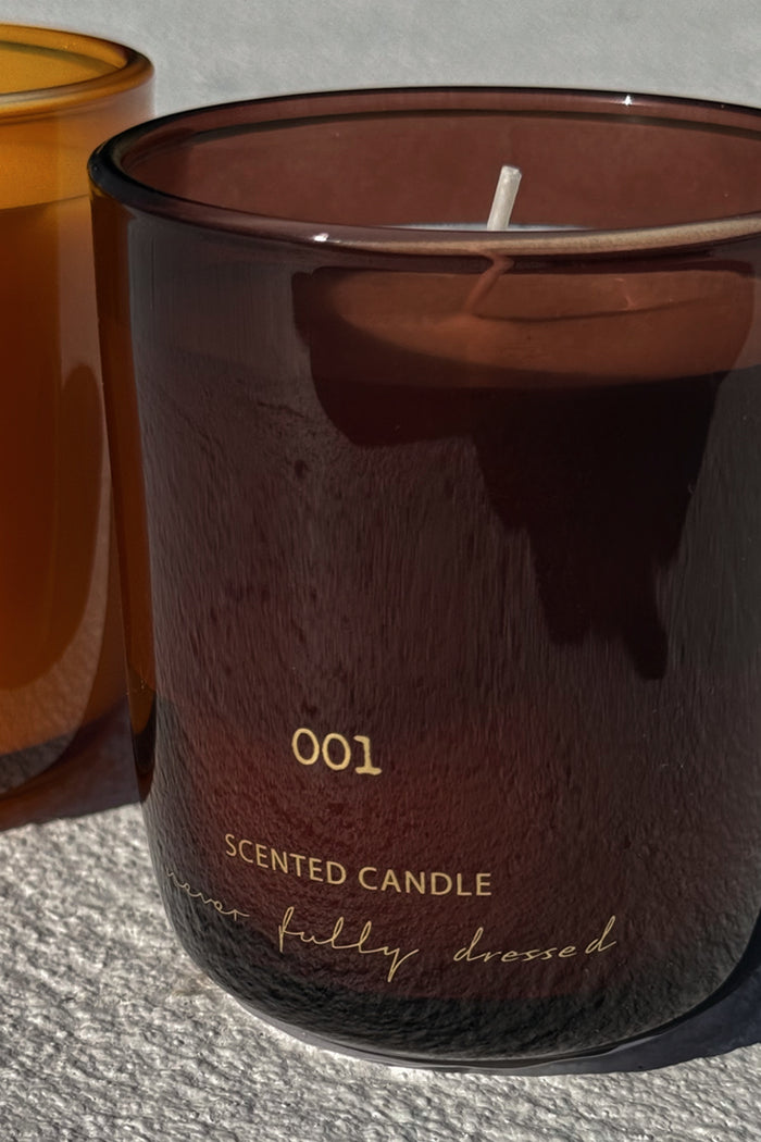 NFD Eau De Parfum Candle 001 in a burgundy glass jar with gold lettering on a sunlit stone surface.
