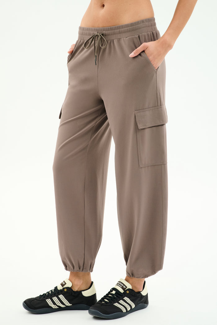 A person wears SPLITS59’s Nico Airweight Cargo Pant in Lentil, featuring a drawstring waist and side pockets, paired with black athletic shoes against a white background—ideal for workouts or casual outings.