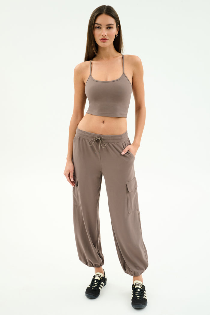 A woman stands against a white background, wearing SPLITS59's Nico Airweight Cargo Pant in Lentil with pockets, styled like joggers, a taupe crop top, and black sneakers.