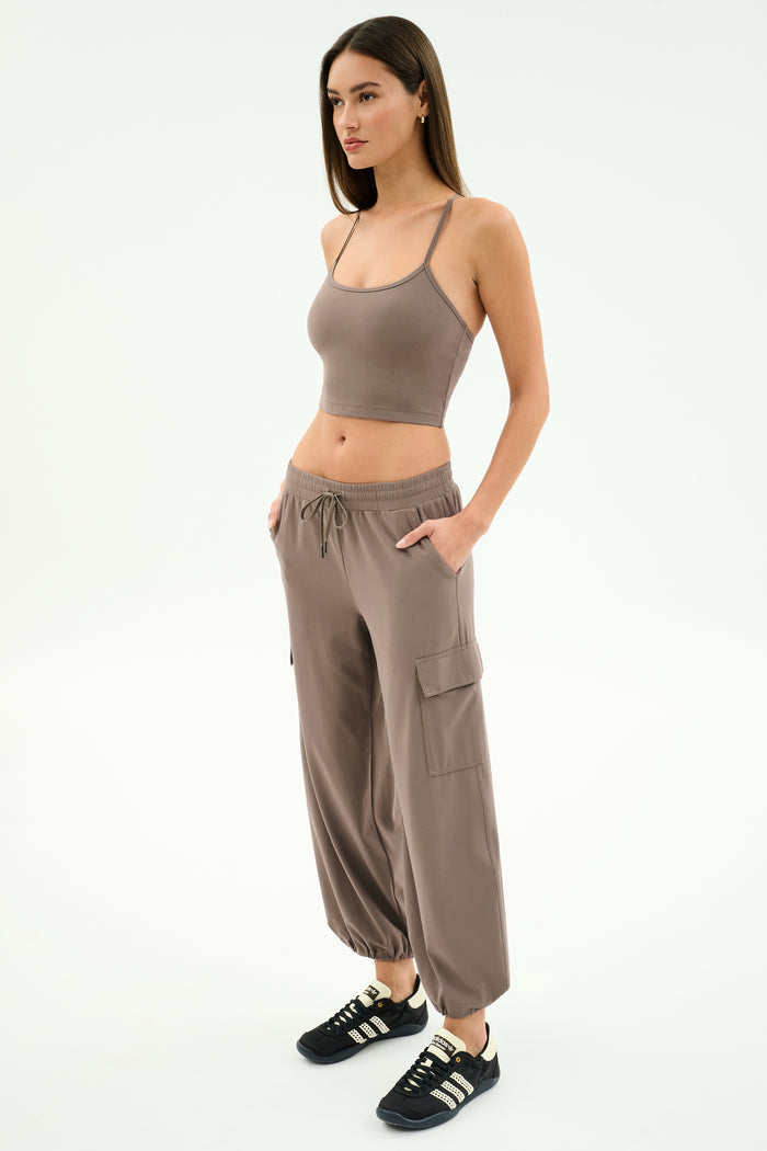 A woman stands in a studio, hands in pockets, wearing SPLITS59’s Nico Airweight Cargo Pant in Lentil with a taupe crop top and black sneakers, perfect for lightweight comfort, facing slightly to the right.