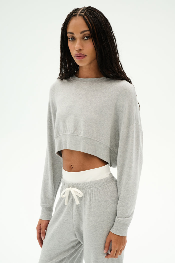 Model wears the SPLITS59 Noah Fleece Crop Sweatshirt in Heather Grey, paired with matching sweatpants featuring white drawstrings, all crafted from soft modal spandex, posed against a plain white background.