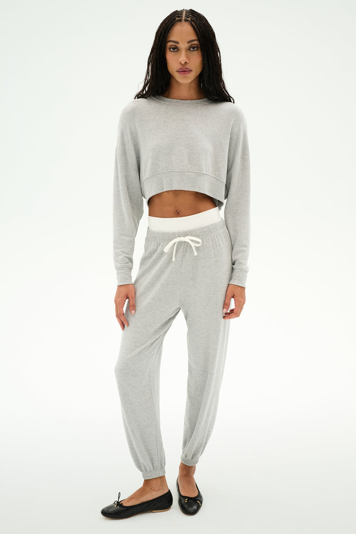 A woman faces forward wearing the SPLITS59 Noah Fleece Crop Sweatshirt in Heather Grey, styled with matching joggers featuring a white drawstring and black flats.