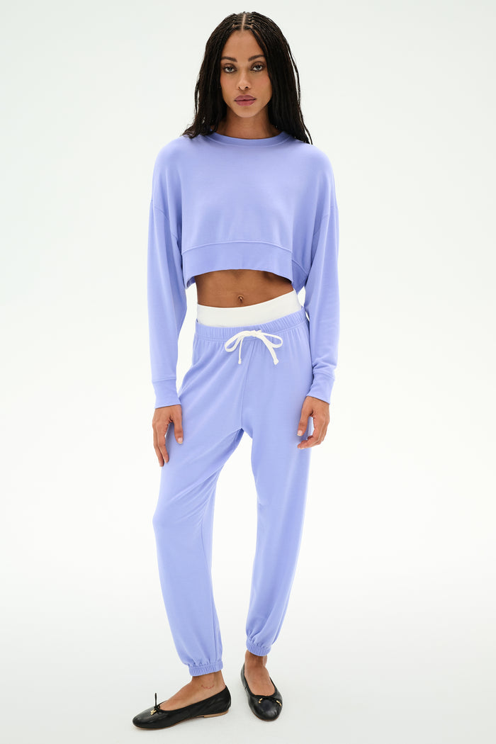 A person wears the SPLITS59 Noah Fleece Crop Sweatshirt in Purple Haze with matching jogger pants, made from a stretchy spandex blend, paired with black ballet flats against a plain white background.
