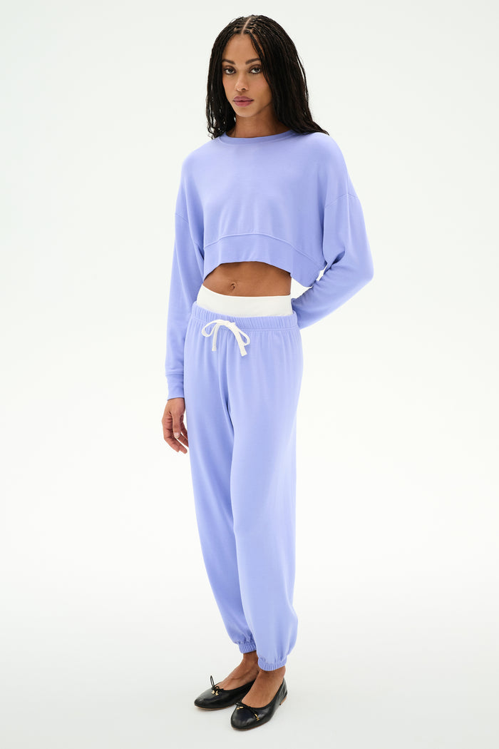 A person stands against a white background wearing the SPLITS59 Noah Fleece Crop Sweatshirt in Purple Haze with matching sweatpants, both made from stretchy material, styled with black ballet flats.