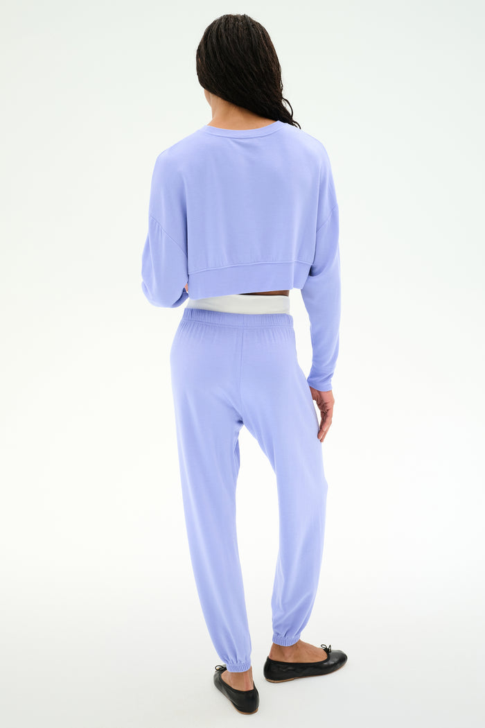 A person faces away from the camera, wearing the SPLITS59 Noah Fleece Crop Sweatshirt in Purple Haze with matching pants and black flats.