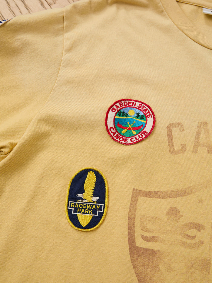The FOUND Patchwork Camp Tee is a yellow unisex relaxed-fit tee featuring vintage-style embroidered patches: a round "Garden State Canoe Club" and an oval "Raceway Park" patch with an eagle graphic.