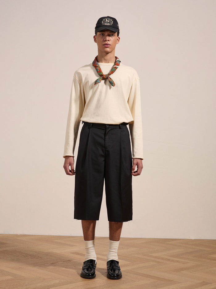 A man wearing a beige long-sleeve shirt, black knee-length FOUND Pleated Trouser Shorts with an adjustable waist, a plaid neck scarf, a black baseball cap, and black dress shoes stands on a wooden floor against a plain background.