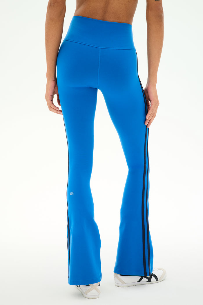 A person is shown from the back against a plain background wearing SPLITS59′s Raquel High Waist Supplex Flare pants in Classic Blue/Indigo, featuring black side stripes and paired with white sneakers. These high-waisted leggings offer 4-way stretch for optimal comfort and flexibility.