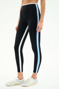 A person models the SPLITS59 Sam High Waist Rigor 7/8 leggings in Black/Skylight, featuring vertical white and blue stripes, paired with white and silver athletic shoes against a plain background.