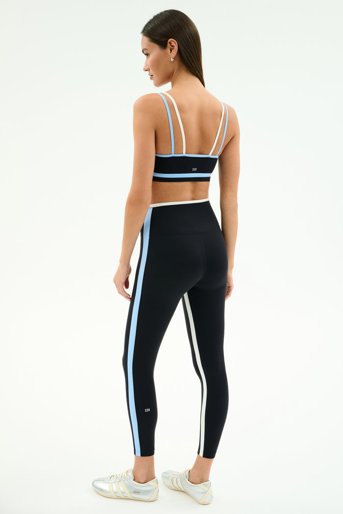 A woman stands sideways on a white background, wearing SPLITS59's Sam High Waist Rigor 7/8 Leggings in Black/Skylight with white and blue side stripes, paired with a matching black run tight sports bra.