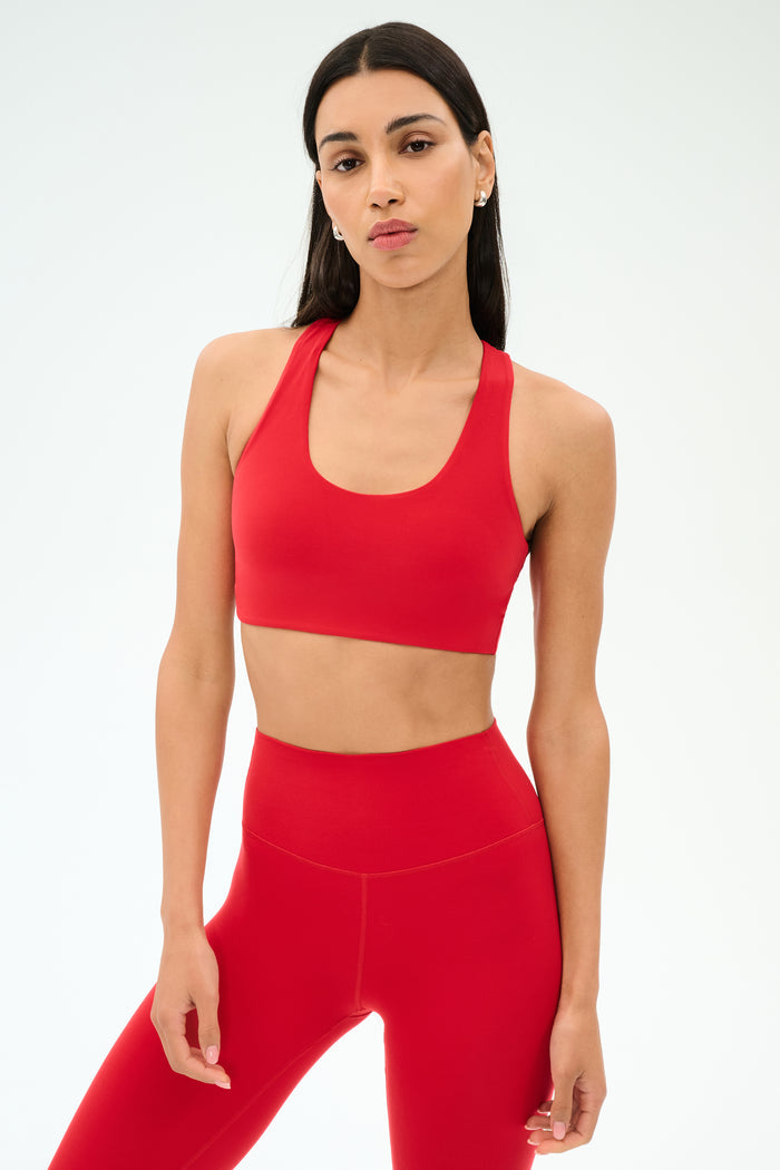 A woman with long dark hair wears the SPLITS59 Sara Airweight Bra in Pirate Red and matching high-waisted leggings against a plain light background—ideal for hot yoga or Pilates.