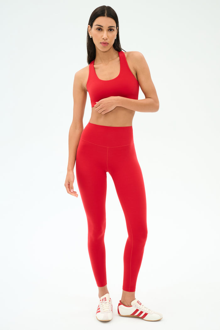 A woman stands against a light background wearing the SPLITS59 Sara Airweight Bra in Pirate Red with matching leggings and white sneakers with red stripes—perfect for Pilates or hot yoga.