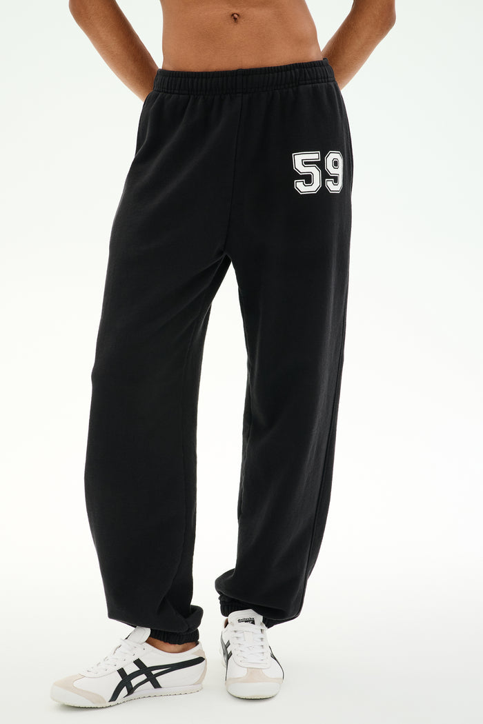 A person is wearing the SPLITS59 Serena French Terry Oversized Sweatpant in black with a “59” on the left thigh, paired with white sneakers featuring black accents, standing against a plain background; these sweatpants are made in Los Angeles.