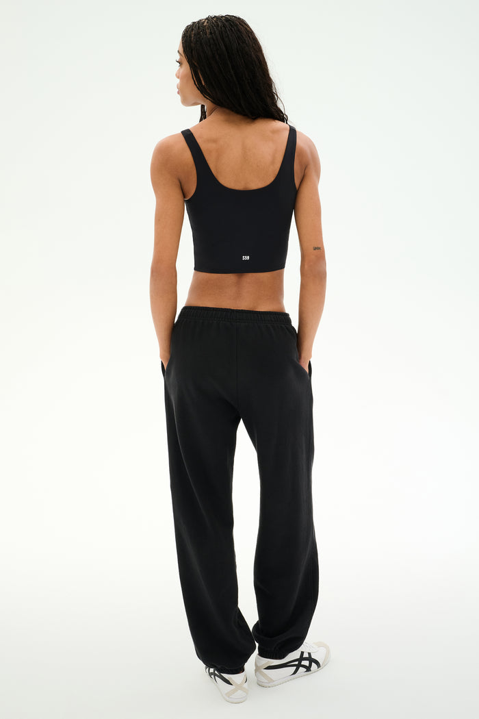 A person stands facing away with hands in pockets against a plain light background, wearing a black crop top, white sneakers, and the SPLITS59 Serena French Terry Oversized Sweatpant in Black/59, made in Los Angeles.