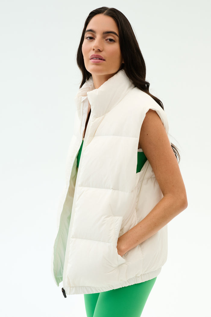 A woman sports the SPLITS59 Summit Nylon Puffer Vest in white over a green outfit, standing with one hand in her pocket against a plain backdrop—perfect lightweight insulation for cool weather workouts.