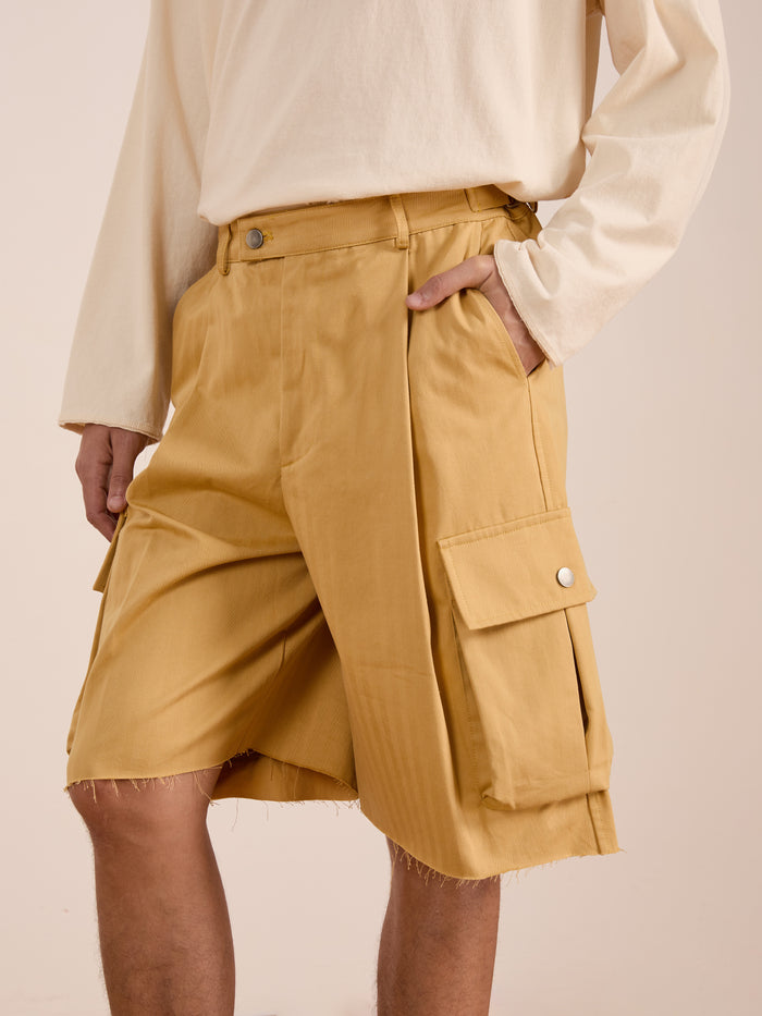 Model in a cream long-sleeve shirt and FOUND Sunlight Twill Cargo Shorts—unisex beige cargos with frayed hems, large side pockets, and subtle herringbone twill texture—stands with one hand in a pocket.